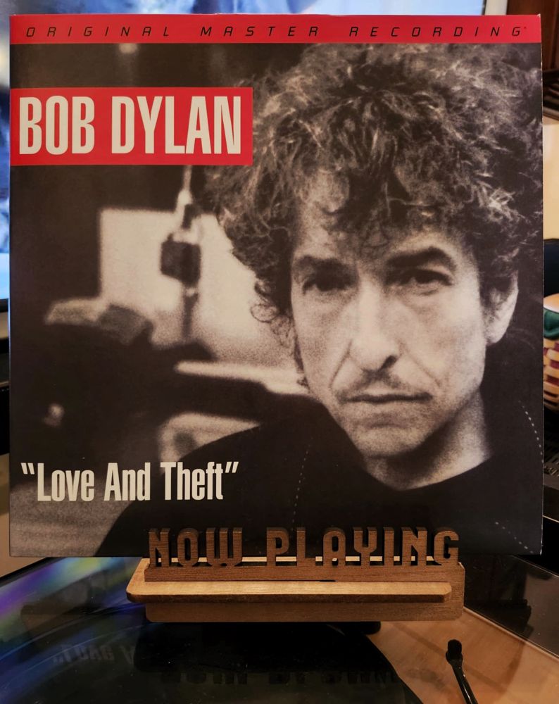 The front cover of the LP record “Love And Theft” by Bob Dylan sits on a wooden “now playing” record display stand placed on top of the turntable that’s playing the album.
A muted television plays in the background.