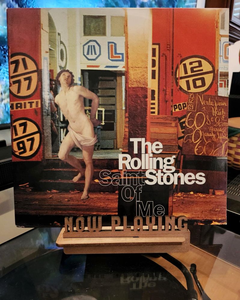 The front cover of the 12” single “Saint Of Me” by The Rolling Stones sits on a wooden “now playing” record display stand placed on top of the turntable that’s playing the album.
A muted television plays in the background.