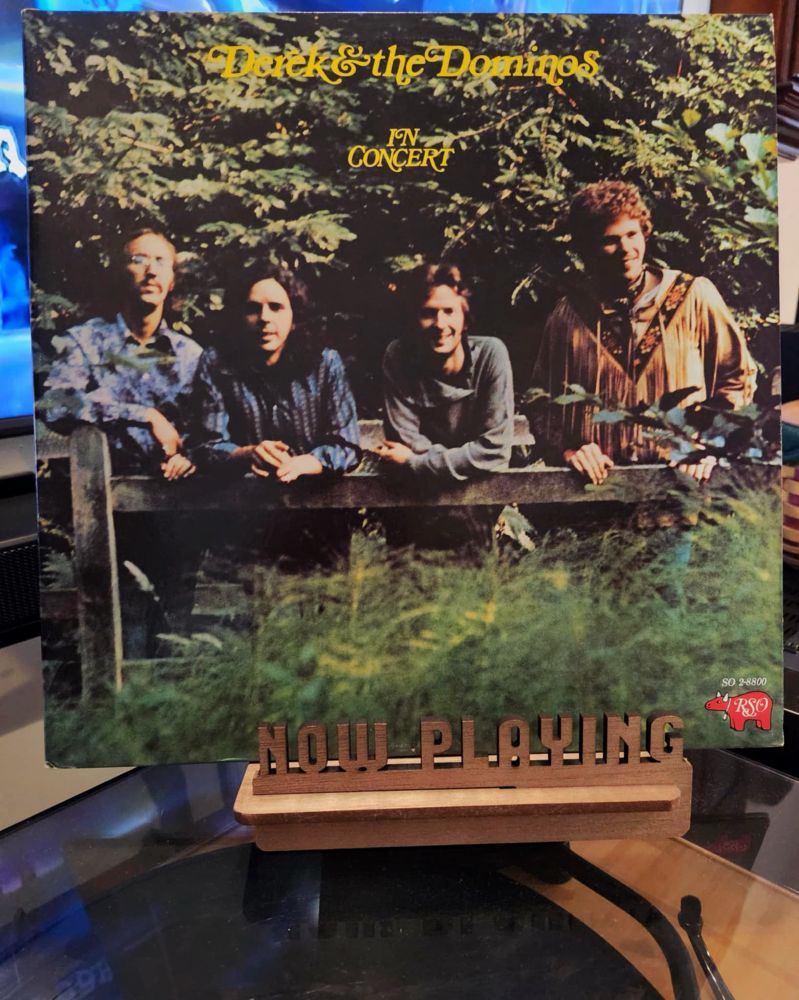 The front cover of the 2-LP live album “In Concert” by Derek & The Dominos sits on a wooden “now playing” record display stand placed on top of the turntable that’s playing the album. A muted television plays in the background.