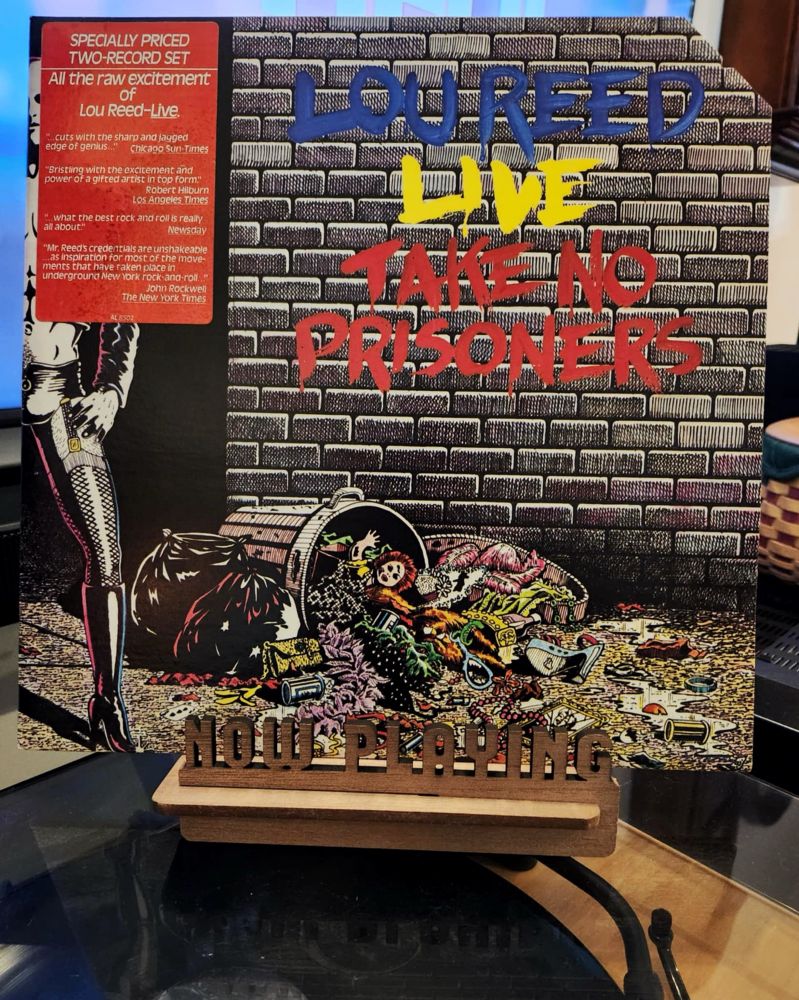The front cover of the 2-LP live album “Take No Prisoners” by Lou Reed sits on a wooden “now playing” record display stand placed on top of the turntable that’s playing the album.
A muted television plays in the background.