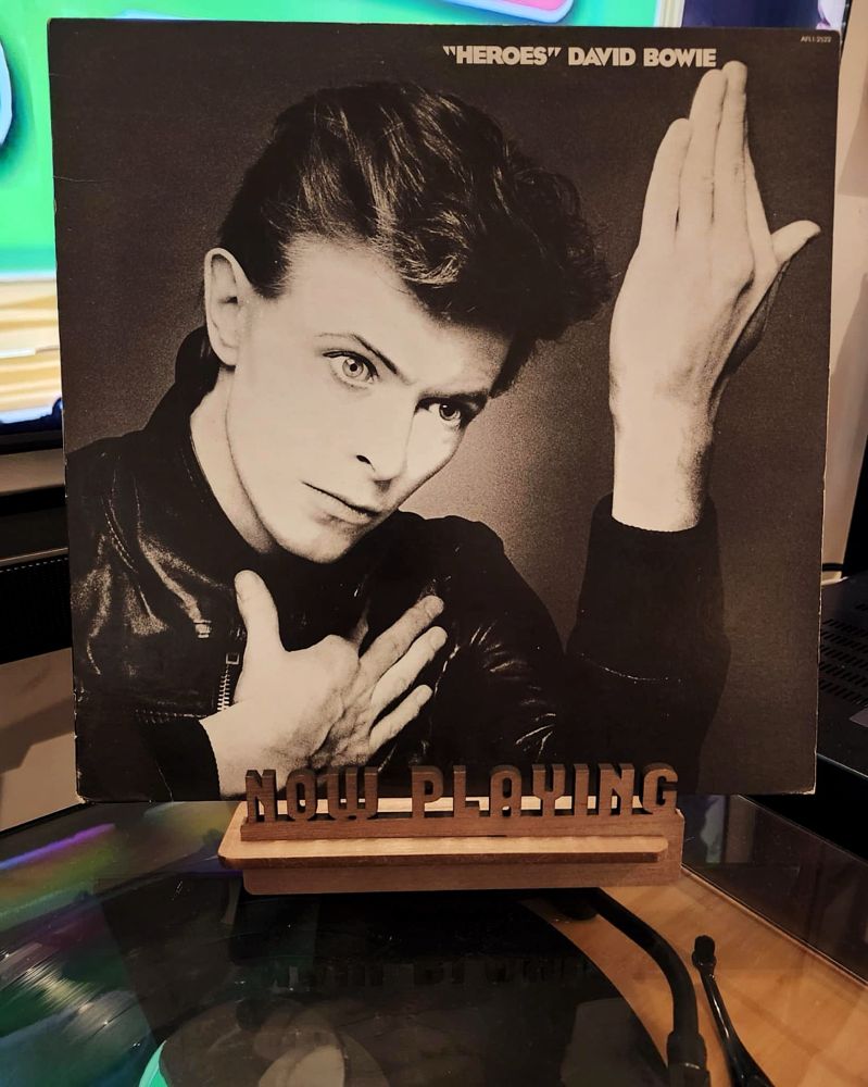 The front cover of the 1977 LP record “Heroes” by David Bowie sits on a wooden “now playing” record display stand placed on top of the turntable that’s playing the album.
A muted television plays in the background.