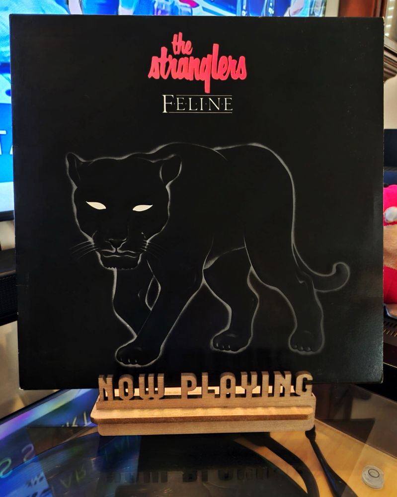 The front cover of the 1982 LP record “Feline” by The Stranglers sits on a wooden “now playing” record display stand placed on top of the turntable that’s playing the album. A muted television plays in the background.