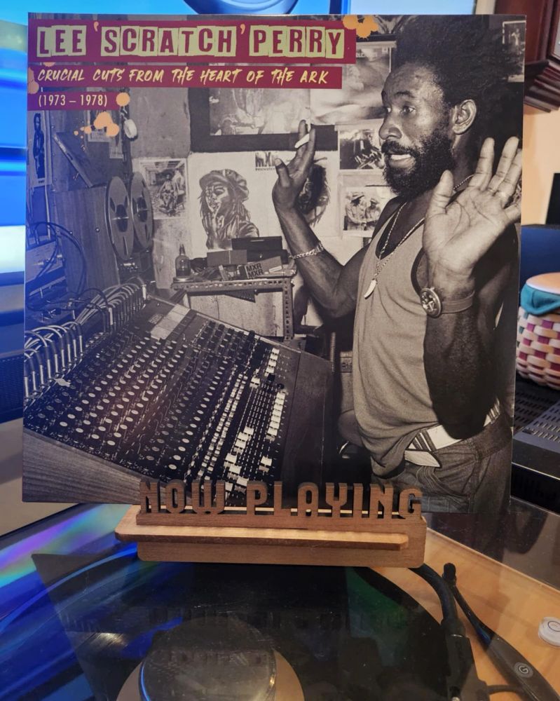 The front cover of the LP record “Crucial Cuts From The Heart of The Ark (1973-1978)” by Lee “Scratch” Perry sits on a wooden “now playing” record display stand placed on top of the turntable that’s playing the album.
A muted television plays in the background.