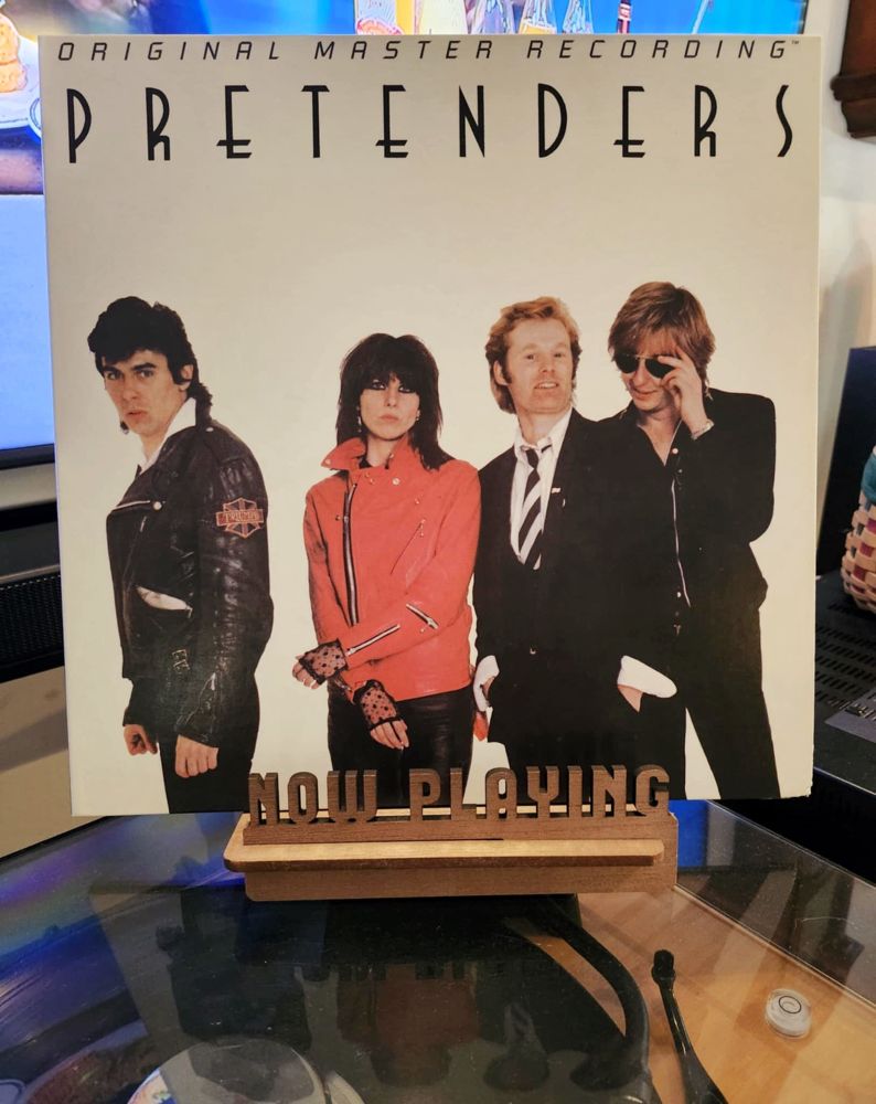 The front cover of the Pretenders’ self-titled debut sits on a wooden “now playing” record display stand placed on top of the turntable that’s playing the album. A muted television plays in the background.