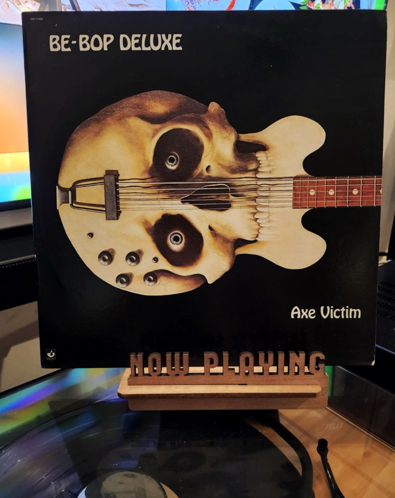 The front cover of the LP record “Axe Victim” by Be-Bop Deluxe sits on a wooden “now playing” record display stand placed on top of the turntable that’s playing the album.
A muted television plays in the background.