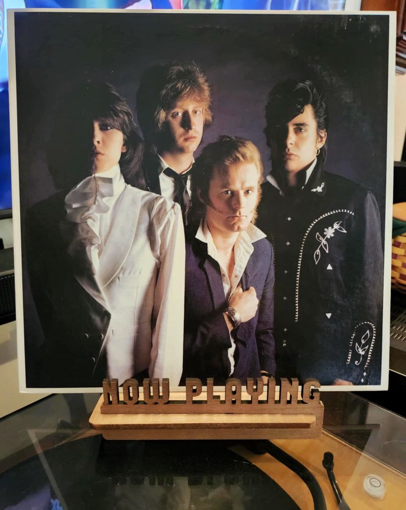 The front cover of the LP record “Pretenders II” sits on a wooden “now playing” record display stand placed on top of the turntable that’s playing the album. A muted television plays in the background.
