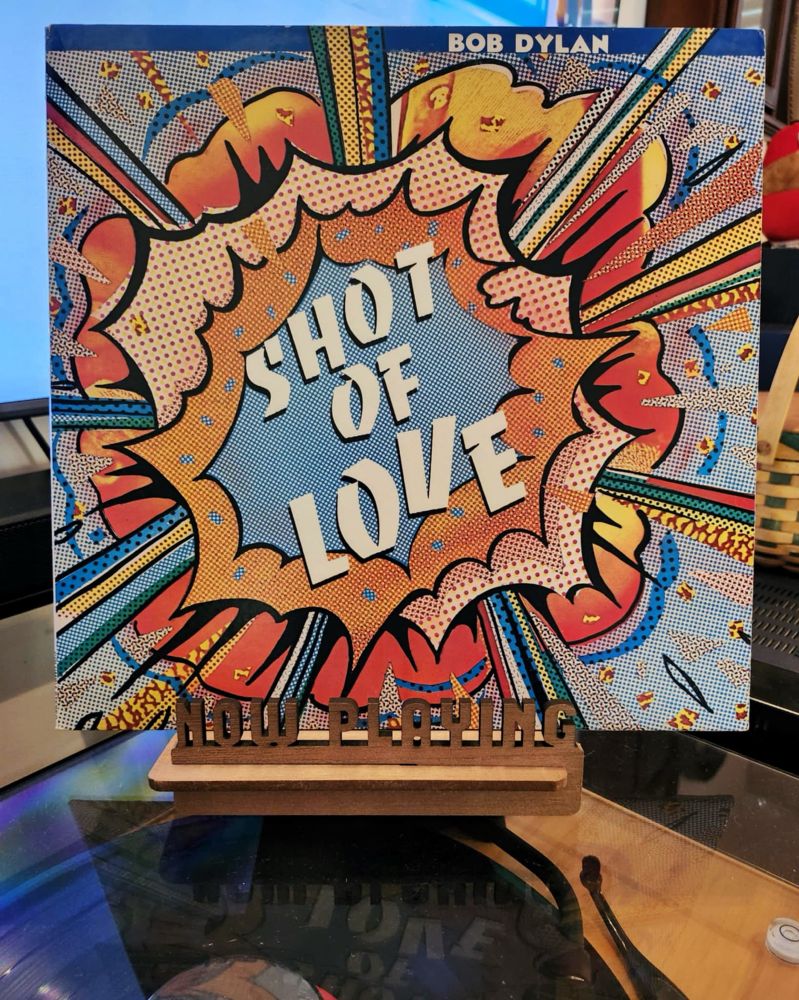 The front cover of the 1981 LP record “Shot Of Love” by Bob Dylan sits on a wooden “now playing” record display stand placed on top of the turntable that’s playing the album.
A muted television plays in the background.