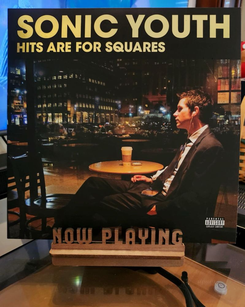 The front cover of the 2-LP vinyl compilation from 2024, “Hits Are For Squares” by Sonic Youth sits on a wooden “now playing” record display stand placed on top of the turntable that’s playing the album. A muted television plays in the background.