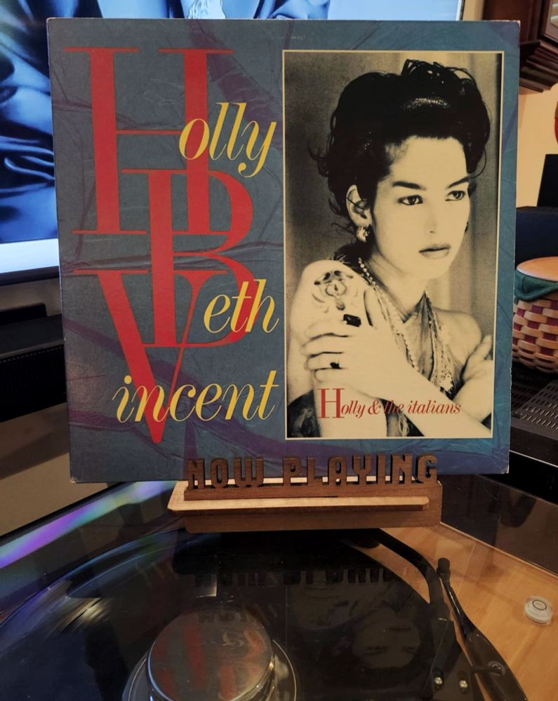 The front cover of the 1982 LP record “Holly And The Italians” by Holly Beth Vincent sits on a wooden “now playing” record display stand placed on top of the turntable that’s playing the album.
A muted television plays in the background.