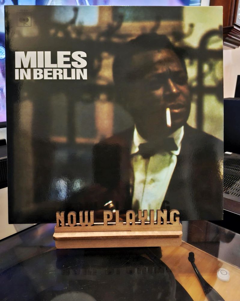 The front cover of the LP record “Miles In Berlin” by Miles Davis sits on a wooden “now playing” record display stand placed on top of the turntable that’s playing the album. A muted television plays in the background.
