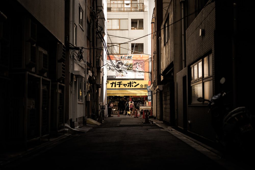 narrow alleyway between tall buildings with a storefront at the end featuring a yellow sign with Japanese characters, surrounded by people and orange cones, with a mix of old and new architecture and a complex network of wires overhead. *) Alt text generated by AI