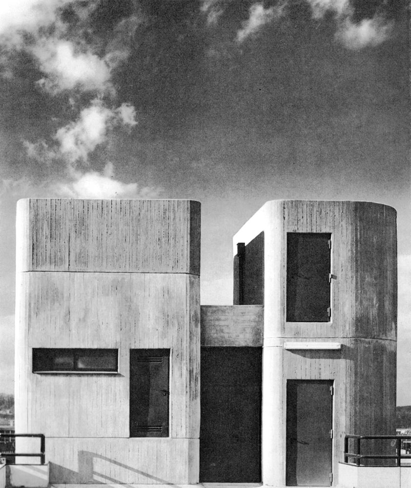 black and white photograph of a concrete building with two cylindrical towers, one with a rectangular window on the left side and the other with a rectangular window on the right side, set against a cloudy sky *) Alt text generated by AI