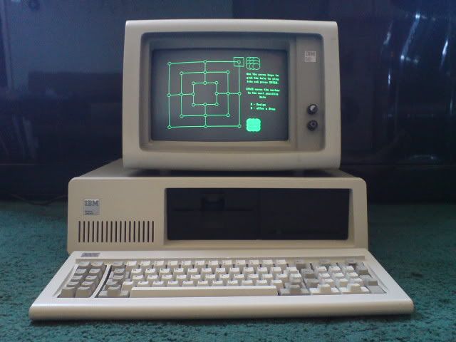 An IBM computer with a beige monitor, system unit, and keyboard, displaying a green graphic and text on the screen, placed on a green carpeted floor. *) Alt text generated by AI