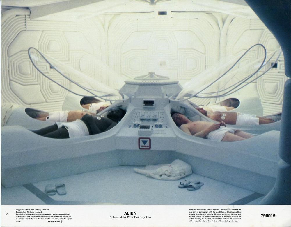 Four people lying on white pods in a white chamber, attached to curved glass tubes, with a central control panel, in a futuristic medical bay, as seen in the 1979 film Alien. *) Alt text generated by AI