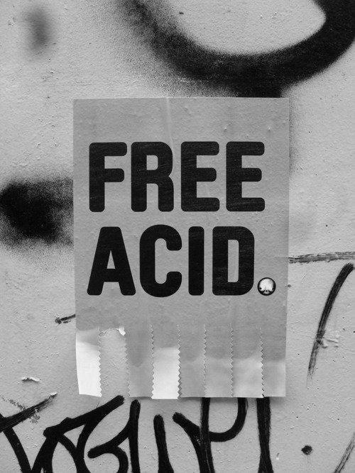 A black and white image of a torn piece of paper stuck on a wall with the words "FREE ACID" printed in large, bold, black letters, surrounded by scattered black graffiti on a gray concrete background. *) Alt text generated by AI