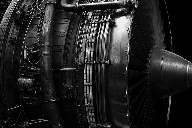 A detailed black-and-white close-up of a large jet engine, showcasing its complex inner mechanisms, with a prominent turbine fan, numerous pipes, and wires against a dark background, highlighting the engine's intricate metal components and reflective surfaces. *) Alt text generated by AI