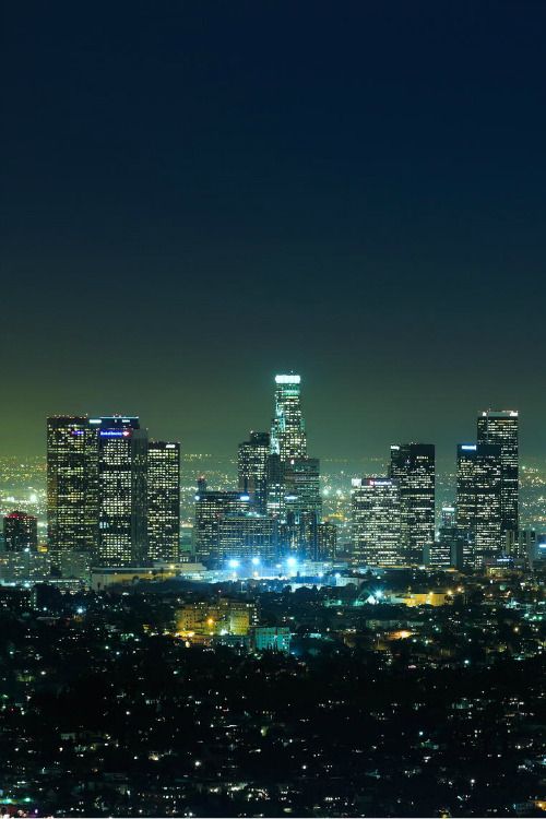 A nighttime cityscape with a cluster of illuminated skyscrapers in the center, surrounded by a densely populated urban area with numerous lights, set against a dark blue and green gradient night sky. *) Alt text generated by AI