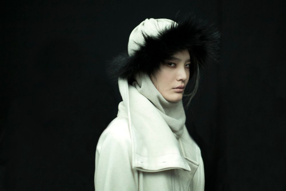 Woman wearing a white parka with a fur-lined hood and a matching turtleneck sweater against a dark background, gazing to the right. *) Alt text generated by AI