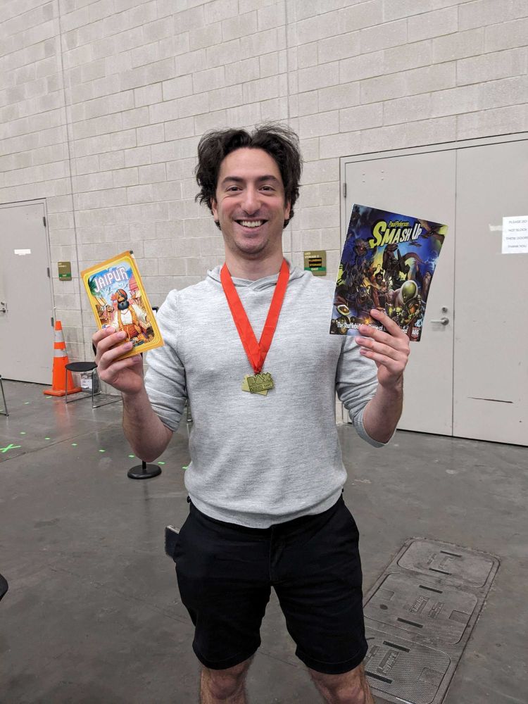 Attendee of PAX East 2025 posing with board games (Smash Up and Jaipur) and two medals