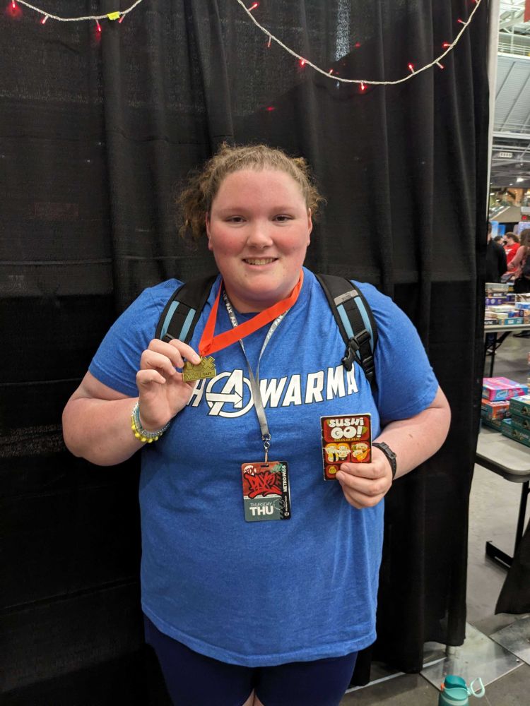 Attendee of PAX East 2025 posing with board game (Sushi Go) and medal