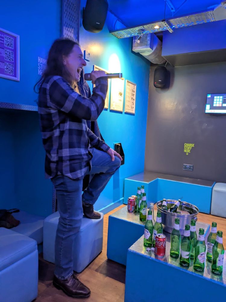 And long haired individual, wearing blue denim and a blue shirt, holding a microphone in a very blue karaoke venue. The individual is clearly making a lot of noise and singing “passionately”. 