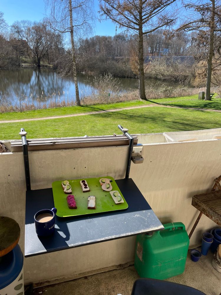 Hjemmelavet smørrebrød på spækbræt på klapbord på altan, med udsigt til lille sø i flot solskin 