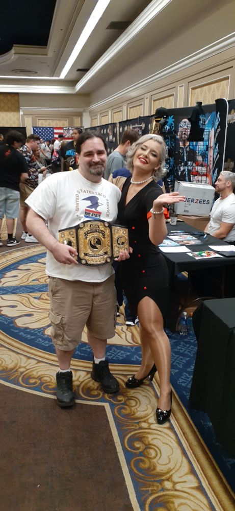 Me, a middle aged white guy with long dark hair in a ponytail and a goatee, holding the AEW women's title belt, posing with the women's champion Toni Storm, a blonde woman in a back pinup dress with red trim and high heels at a wrestling convention in a hotel ballroom.