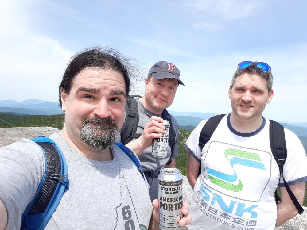 Three smiling middle aged white guys posing at the top of a mountain in New Hampshire, holding NH beers