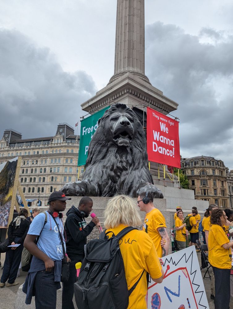 Celebration in Trafalgar Square. One of the lions at the base of Nelson's column is centered and two banners attached behind the lion's head. Red banner, small white text header "they wanna fight" main text "We Wanna Dance". Obscured green banner text but it says "Freedom to Party". Various people in the foreground, many look like they are volunteers as most are wearing yellow T-shirts. 