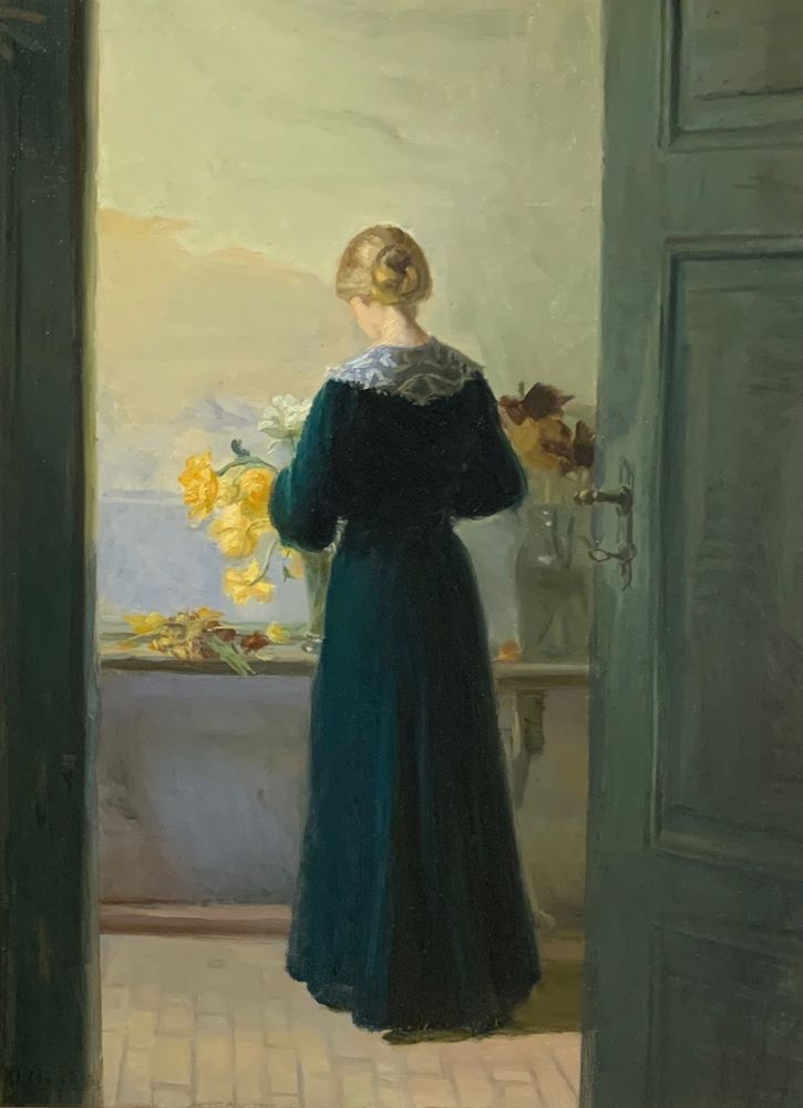 Blonde Frau in dunklem Kleid von hinten. Sie steht vor einer hellen Wand und arrangiert gelbe und weiße Blumen.