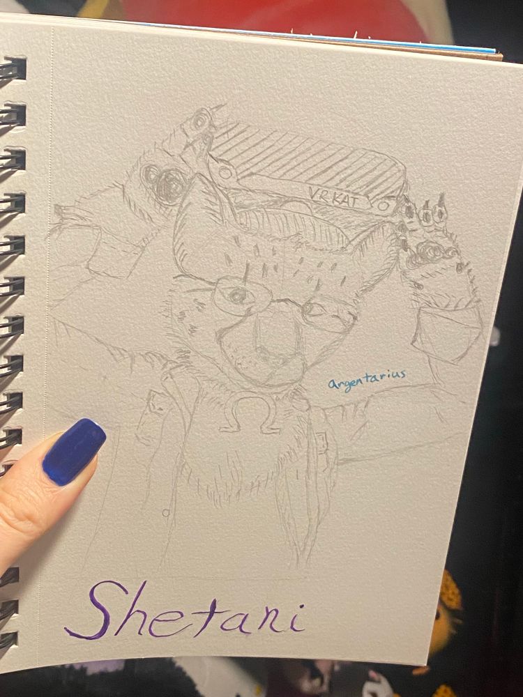 I hold my sketchbook to show the sketched version of Shetani putting on VR goggles that say "VR Kat" on the front. The iconic Omega necklace can be seen. Under the drawing is "Shetani". Over the bicep "Argentarius" (the artist) is written. My thumb nail is painted metallic blue.