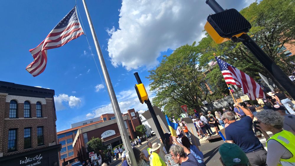 Photo of community members protesting at Court & Main Streets in Doylestown, many holding signs, two American flags flying in the breeze. (Lots of drivers going by, honking in support!)