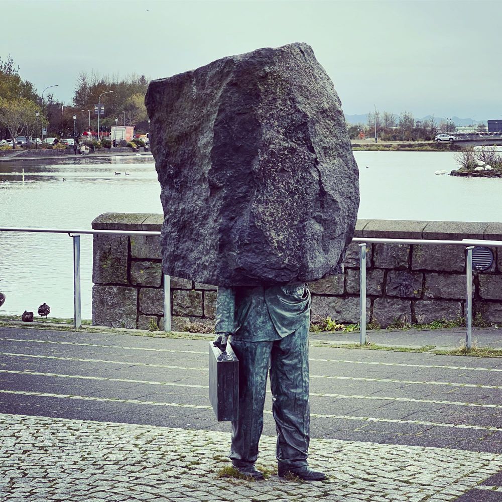 Bronze statue of a man’s legs and hand holding a briefcase with the top half of his body replaced by a boulder. In Reykjavik 