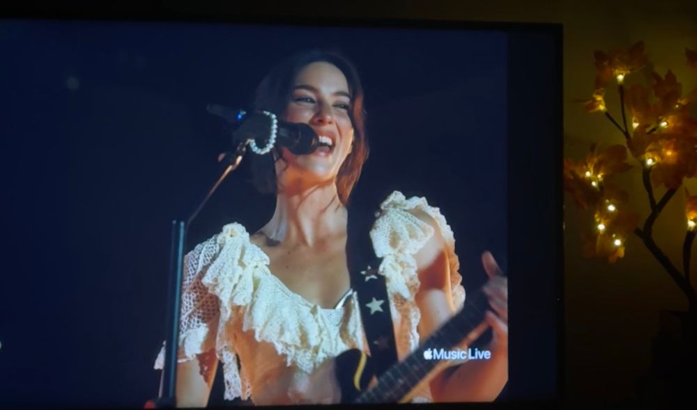photo of gracie abrams from the Apple Production of her performance from Red Rocks on my tv. Gracie is wearing a crocheted white dress with sleeves and holding a guitar with stars on the strap. She’s smiling while singing into a microphone that’s on its stand and has a friendship bracelet around it. to the side of my tv you can see a small tree with branches lit up with orange lights as fall decor. The Apple logo Music Live is in the bottom right corner 
