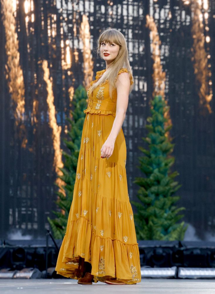 taylor swift standing on stage at the eras tour wearing one of the original dark burnt orange/gold evermore dresses. she is smiling out at the crowd and you can see trees and stage effects lit up behind her.