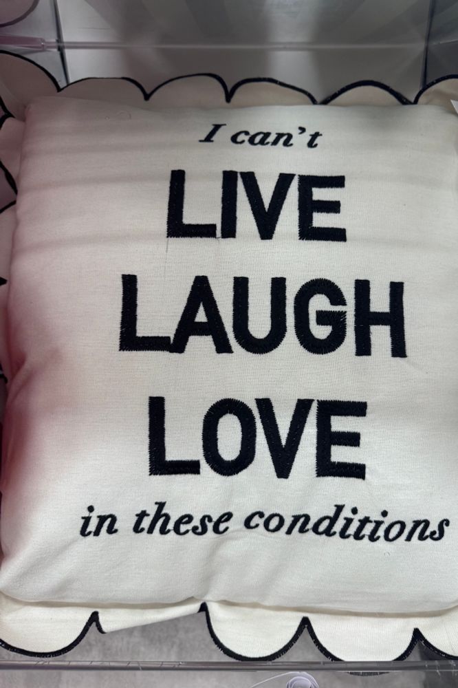 picture of a beige pillow with black writing that says “i can’t live laugh love in these conditions”. the sides of the pillow have a scallop design outlined in black