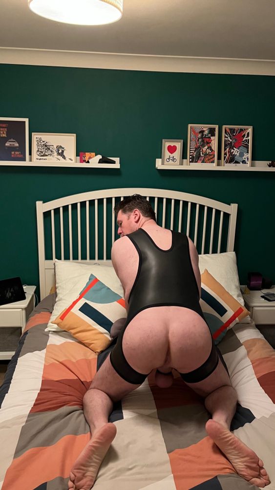 40 year old male on a bed in a neoprene singlet open at the rear and a black chastity cage looking back over his shoulder.