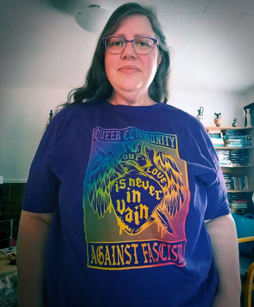 Dee, a fat white woman wearing glasses and with long brown and gray hair, is wearing a purple t-shirt emblazoned with a Pepper Raccoon design on it. In rainbow wash it shows a biological heart, sword and wings, and the words 'our love is never in vain'. Around the exterior of the piece are the words, fully capitalised: 'Queer Community Against Fascists'. Dee is standing in their lounge and some bookcases and greek vases can be seen in the background. 