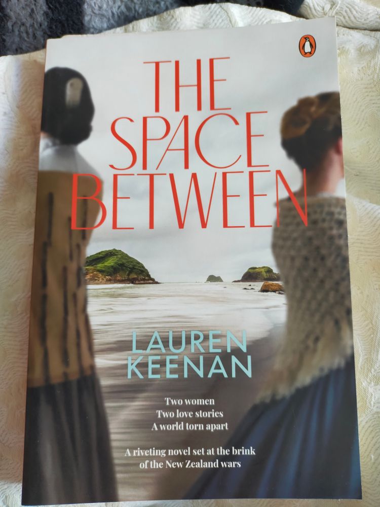 Book cover of 'The Space Between' by Lauren Keenan. Illustrated with side views of a Māori wahine and a British woman facing away from one another (in their own mid 19th century clothing) and the Taranaki coast between them. Text reads "Two women / Two love stories / A world torn apart. A riveting novel set on the brink of the New Zealand wars" 