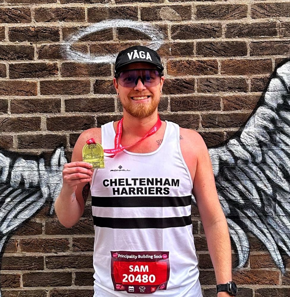 Photo of Sam holding up his Cardiff Half Marathon finishers medal in front a wall painted with angel wings