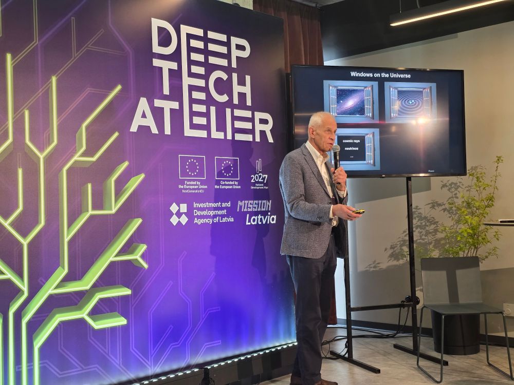 A man in a grey blazer stands on an indoor stage, holding a microphone and presentation remote. To his right, a large monitor displays a slide titled “Windows on the Universe” with space-themed images. To his left, a purple conference backdrop reads “DEEP TECH ATELIER” and features a neon-style circuit-tree graphic alongside EU and “Mission Latvia” logos. An empty chair and a small potted plant sit in the background.