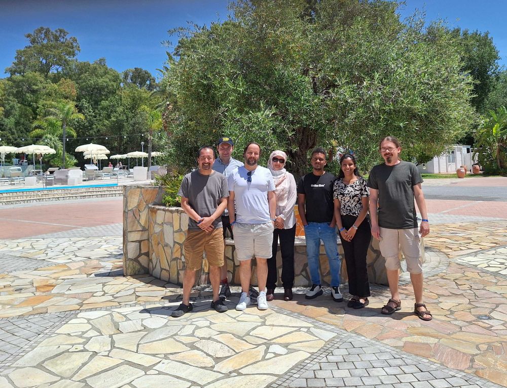 Astrocent team members posing together during the DarkSide-20k summer collaboration meeting in Sardinia, June 2025. The group stands in front of a tree with a pool area and greenery in the background.