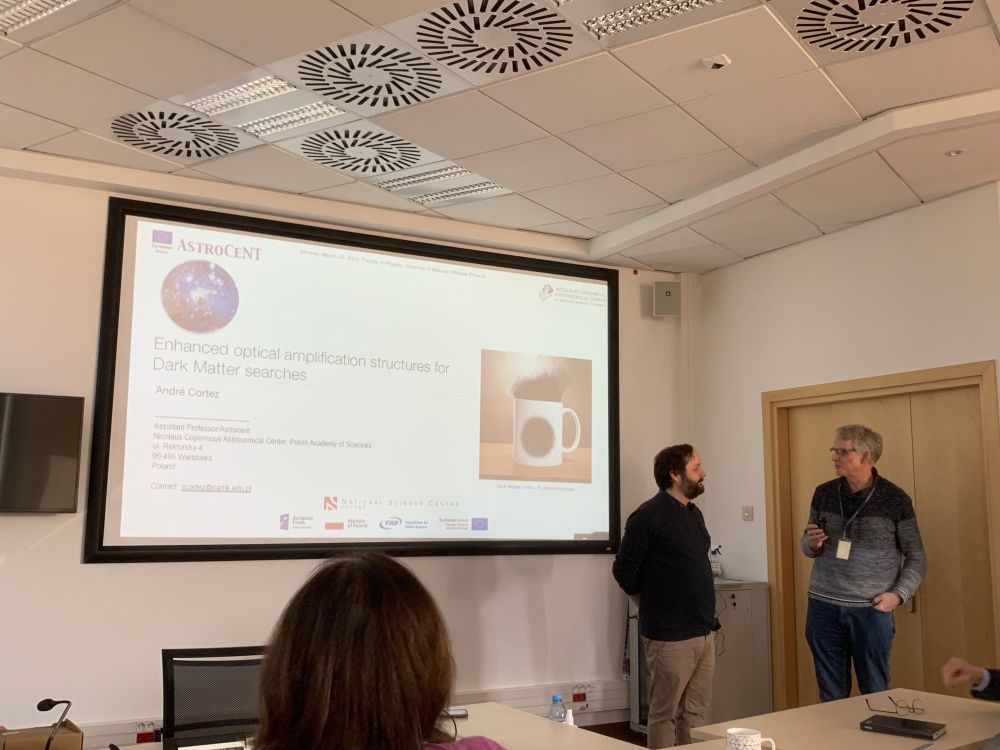  A researcher talking with a colleague in front of a projected slide titled "Enhanced optical amplification structures for Dark Matter searches" during a seminar.