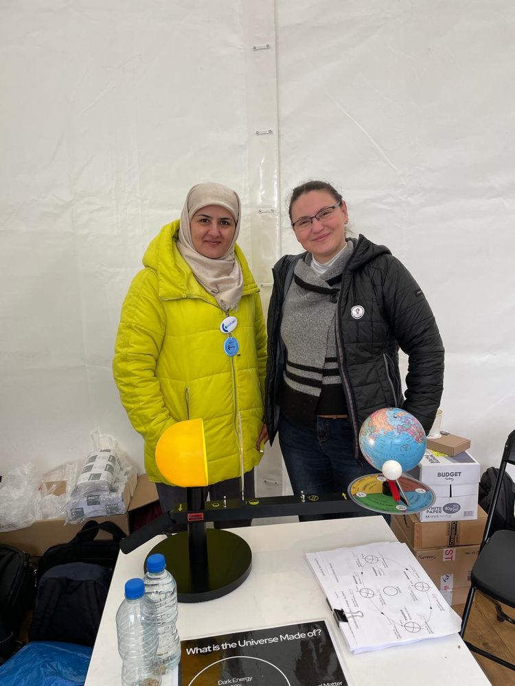 Two women stand side by side, smiling next to their planetary-motion model at the Science Picnic stall.