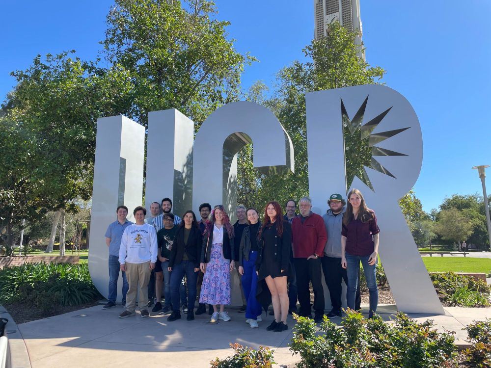 A group of people poses in front of a large UCR (University of California, Riverside) sign outdoors. The group consists of men and women dressed casually and professionally. Behind them, there are trees, bushes, and the UCR bell tower under a clear blue sky.