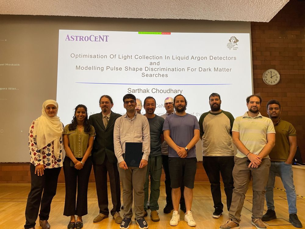 A group of ten people standing in front of a large presentation screen displaying the PhD thesis title. The PhD candidate stands at the center holding a diploma folder. Everyone is smiling, and the setting is a lecture hall.