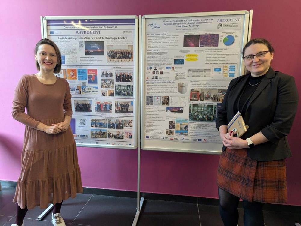 Two women stand next to research posters at the Widening in Horizon Europe event at Warsaw University of Technology. The posters showcase Astrocent's communication and dissemination activities and the DarkWave project on dark matter search technologies. One woman wears a brown dress and white sneakers, while the other wears a black blazer with a plaid skirt and holds a booklet. The background features a pink wall.