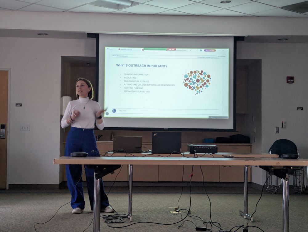 A woman in a white turtleneck and blue pants presents in a conference room, standing in front of a projected slide titled "Why is Outreach Important?" The slide lists key points such as sharing information, educating, and getting funding. She gestures with her hands while speaking. A laptop and projector are set up in front of her.