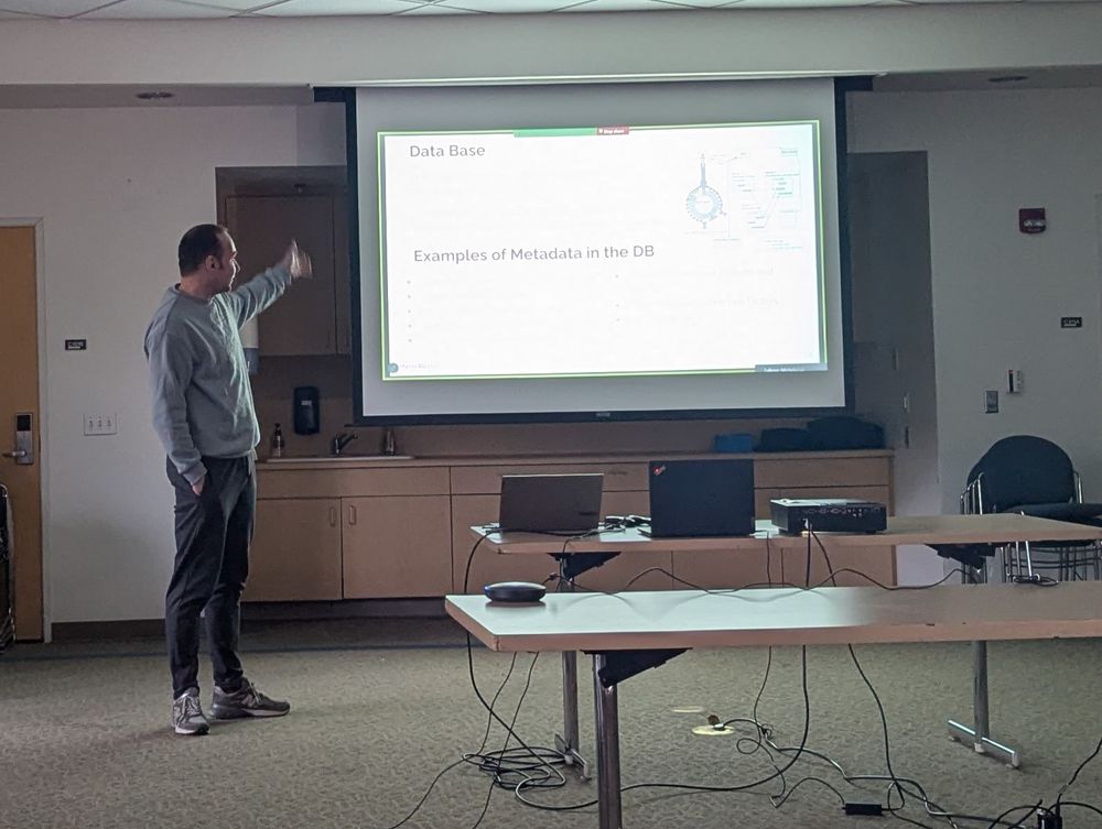 A man in a gray sweater and dark pants stands at the front of a conference room, pointing at a projected slide titled "Data Base" with the subtitle "Examples of Metadata in the DB." The room has a projector, laptops, and a table with cables. The lighting is dim to make the presentation more visible.