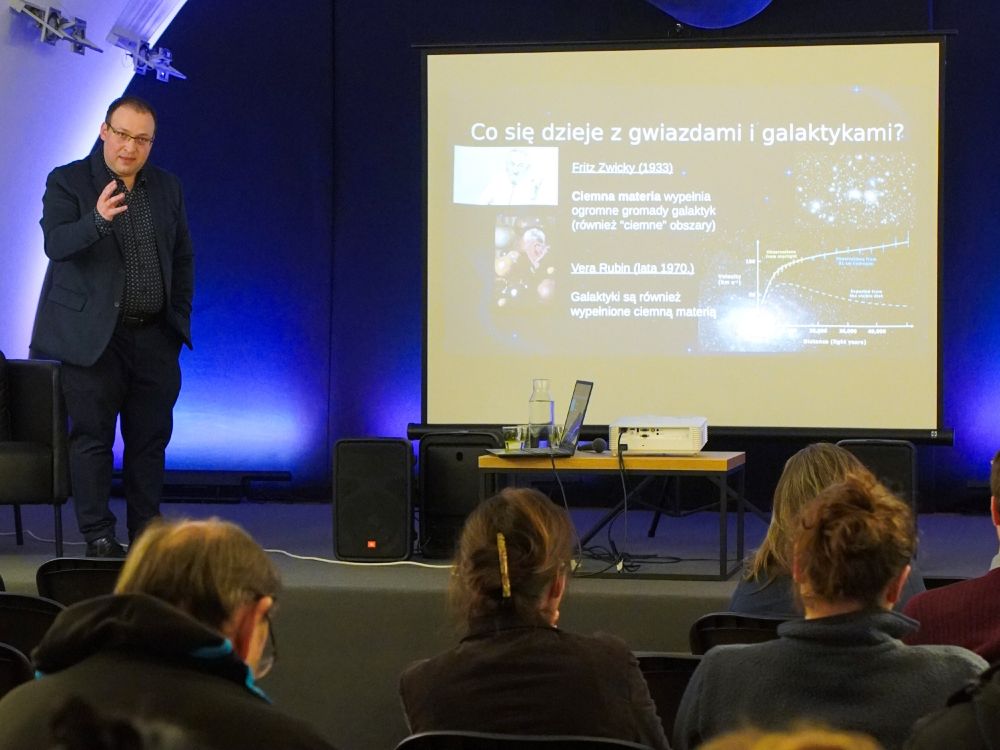 A presenter explains a slide about dark matter in galaxies to an audience during the public lecture.